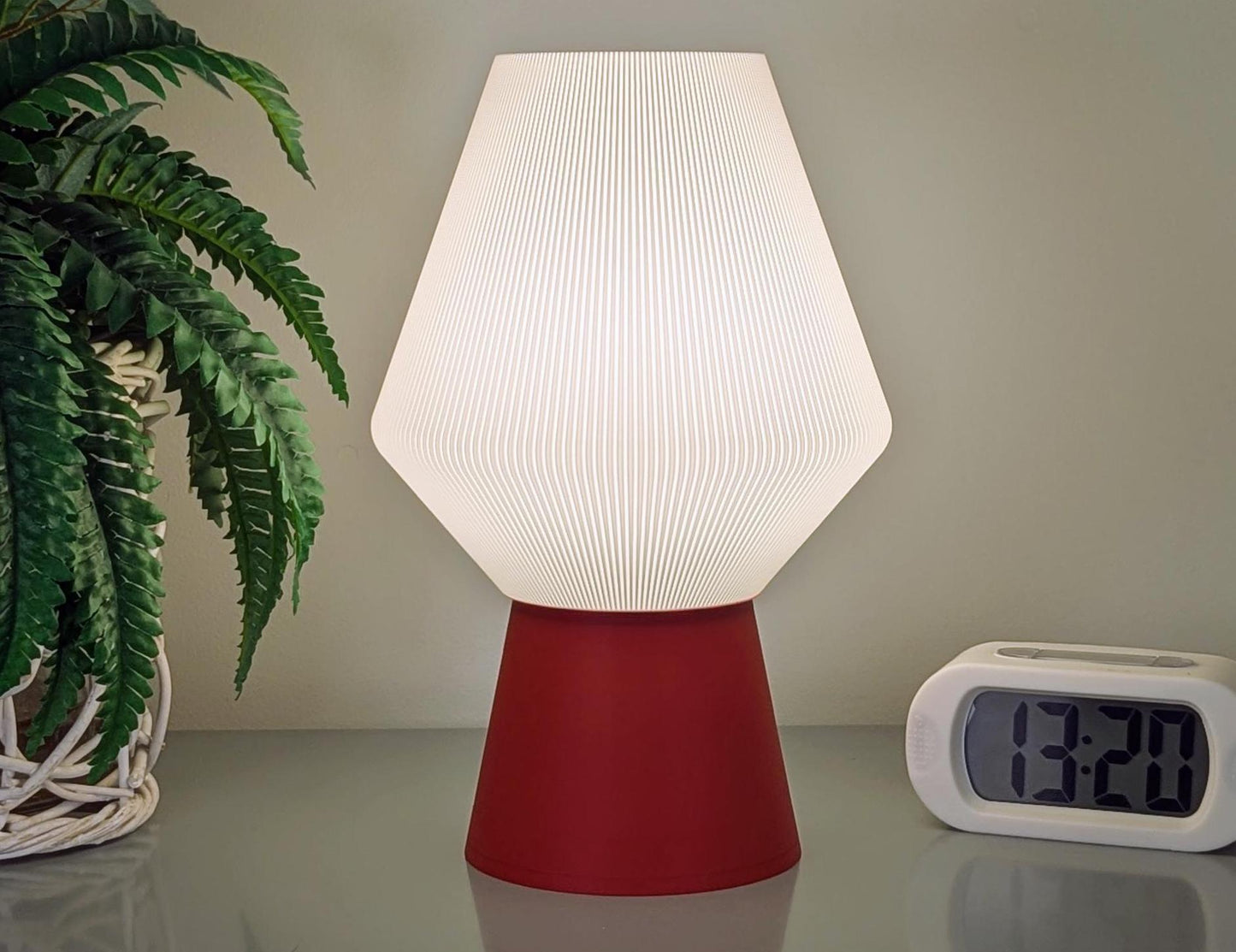 The Inversis Table Lamp by Dryft Home, featuring a white pleated shade and red biodegradable PLA base, sits illuminated on a reflective surface. A digital clock reads 13:20 on the right, while a potted fern in a basket highlights sustainable production on the left.