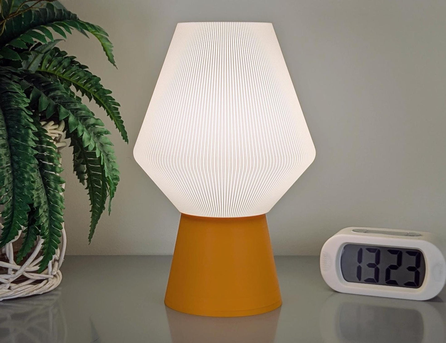 The Inversis Table Lamp by Dryft Home, featuring a white textured shade and orange base crafted from biodegradable PLA, emits a gentle glow. A digital clock showing 13:29 sits to the right with a green potted plant on the left, all resting on a smooth surface.